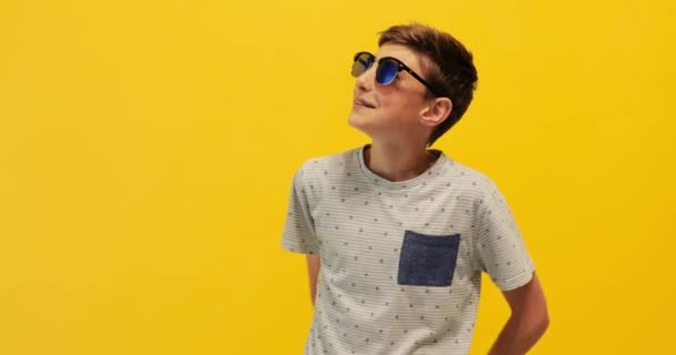 Happy stylový dospívající chlap v slunečních brýlích stojící na žlutém pozadí — Stock video