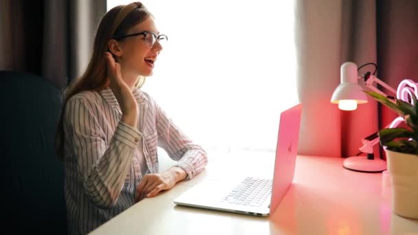 Młoda uśmiechnięta kobieta w okularach komunikująca się z klientem przez filmik z laptopa — Wideo stockowe