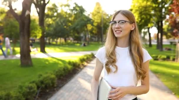 Sorridente bella studentessa che cammina nel parco in una giornata di sole con un computer portatile in mano dopo la lezione — Video Stock