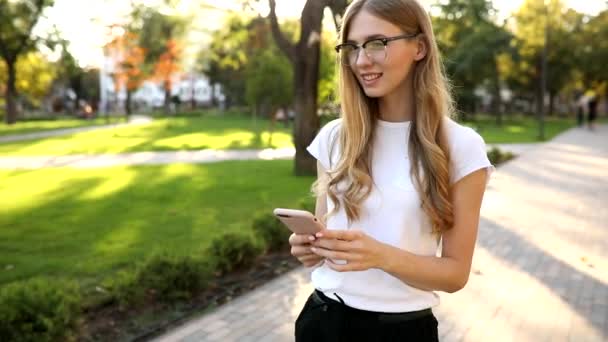 Hermosa joven con cabello rubio, utiliza un teléfono móvil, intercambia mensajes en un teléfono inteligente, al aire libre en un parque — Vídeo de stock