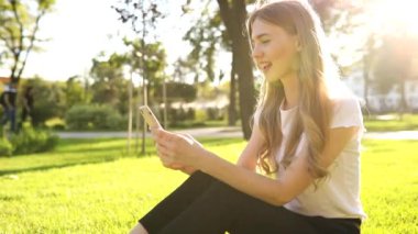 Güzel gülümseyen genç bir kadın bir yaz günü çimlerin üzerinde oturuyor ve dışarıda parkta cep telefonu kullanıyor.
