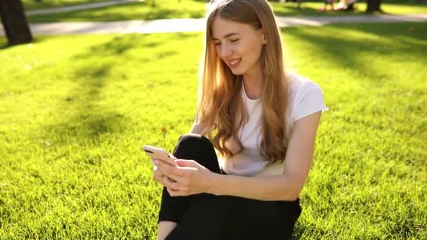 Bella giovane donna sorridente seduta sull'erba in una giornata estiva e utilizzando un telefono cellulare, all'aperto in un parco — Video Stock