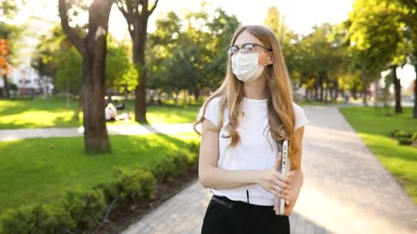 Ung vacker kvinna i en medicinsk skyddsmask i ansiktet, går i parken på en solig dag med en bärbar dator i händerna, coronavirus, epidemi — Stockvideo