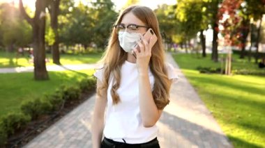 Tıbbi koruyucu maskeli güzel bir kadın şehirde sokakta yürüyor ve bir cep telefonuyla konuşuyor, Coronavirus. 