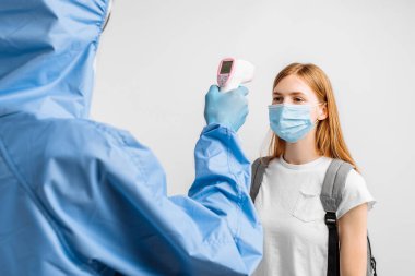 Covid-19 Coronavirus koruyucu tulumlar giyen doktor beyaz bir arka planda genç bir kadının sıcaklığını kontrol etmek için dijital sıcaklık tarama cihazı kullanıyor.