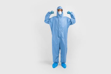 Koronavirüsle savaşmak için koruyucu giysili bir doktor, gözlük, maske ve eldiven takıyor, beyaz arka planda, sağlık hizmetleri konseptinde, pazılarda bir güç hareketi gösteriyor.