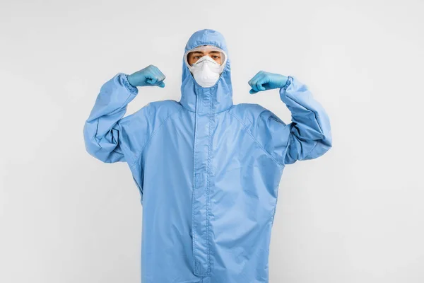 doctor in a protective suit to fight coronavirus, wearing glasses, a mask and gloves shows a gesture of strength on the biceps on a white background, healthcare concept