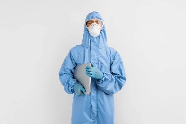 Male doctor in a protective medical suit to protect against coronavirus, in a mask, glasses and gloves, with a laptop on an isolated white background