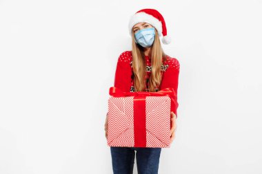 Hediye teslimatı! Noel Baba şapkası ve tıbbi koruyucu maske takan çekici genç bir kadın beyaz arka planda izole edilmiş yeni yıl hediyeleriyle dolu büyük bir hediye kutusu tutuyor.