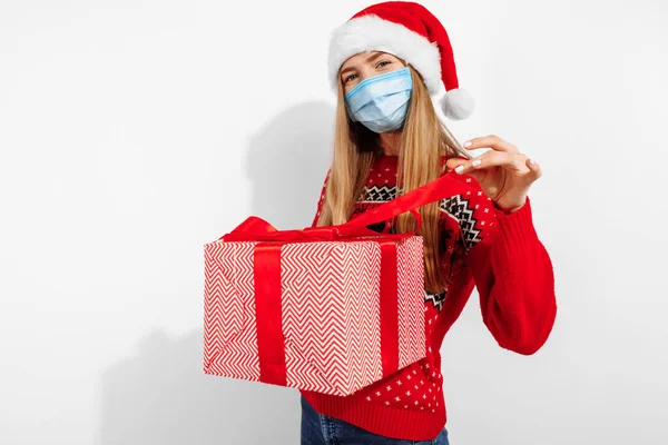 Hediye teslimatı! Noel Baba şapkası ve tıbbi koruyucu maske takan çekici genç bir kadın beyaz arka planda izole edilmiş yeni yıl hediyeleriyle dolu büyük bir hediye kutusu tutuyor.