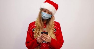 Noel Baba şapkalı, yüzünde tıbbi maske olan gülümseyen sarışın kadın beyaz arka planda bir akıllı telefona bir mesaj yazıyor.