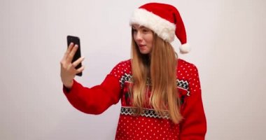 Noel süveteri giymiş, kırmızı Noel Baba şapkalı genç bir kadın Noel arifesinde beyaz bir arka planda akıllı telefondan elini sallıyor.