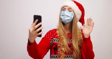 Kırmızı Noel Baba şapkalı, yüzünde tıbbi bir maske olan genç mutlu bir kadın Noel arifesinde beyaz bir arka planda akıllı bir telefondan arama yapıyor.