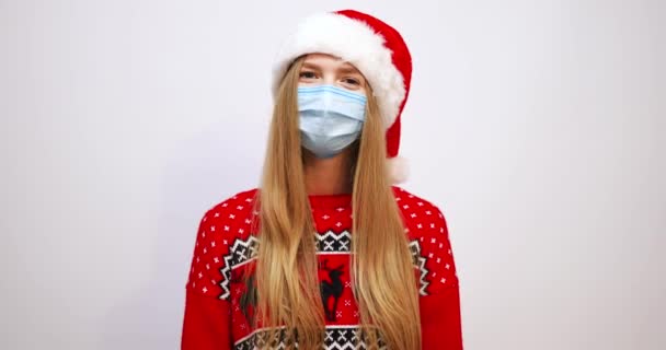 Glad ung jultomte kvinna i jul hatt pekar med pekfingret till medicinsk ansiktsmask skyddad från covid-19 coronavirus virus, på vit bakgrund, nyårsfirande semester — Stockvideo