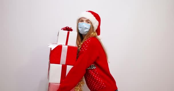 Jovem mulher feliz no Natal chapéu de Papai Noel, em uma máscara médica em seu rosto, com presentes de Natal, em um fundo branco, ajudante de Santas — Vídeo de Stock