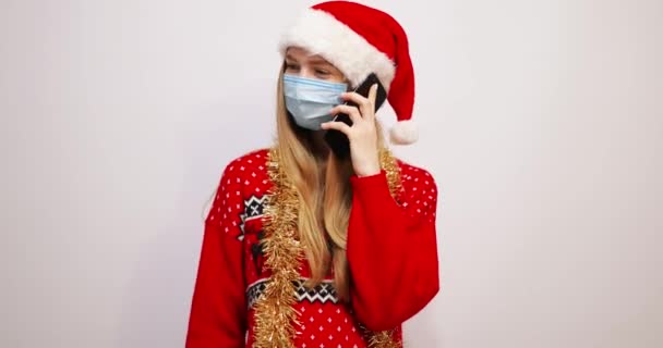白い背景にスマートフォンを持って電話で話している彼女の顔にサンタクロースの帽子と医療用マスクを身に着けている若い幸せなクリスマスの女性 — ストック動画