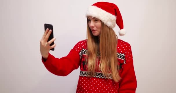 Ung lycklig kvinna i en jultröja och i en röd jultomte hatt, viftar med handen, gör ett videosamtal på en smartphone på julafton, på en vit bakgrund — Stockvideo