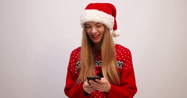 Lächelnde junge Frau mit Weihnachtsmannmütze, benutzt Handy, tippt SMS auf weißem Hintergrund — Stockvideo