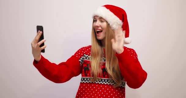 Mulher feliz em uma camisola de Natal e em um chapéu vermelho Papai Noel, acena sua mão, faz uma chamada de vídeo em um smartphone na véspera de Natal, em um fundo branco — Vídeo de Stock