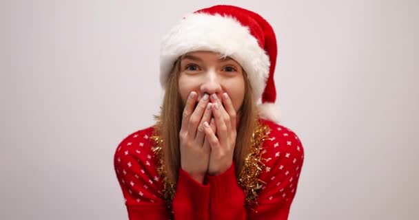 Zszokowana młoda dziewczyna, w świątecznym swetrze i kapeluszu Świętego Mikołaja, patrzy w obiektyw z zaskoczenia na białym tle — Wideo stockowe