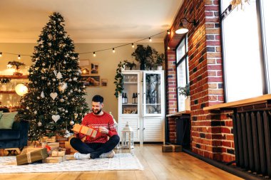 Smiling young man sitting near the Christmas tree, with a Christmas present at home, New Years celebration, Merry Christmas