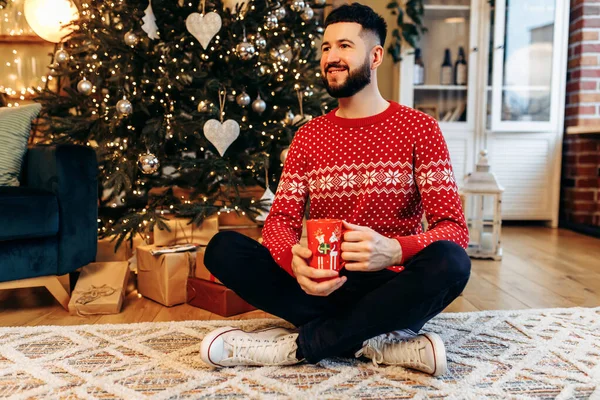 Handsome young man in a Christmas sweater with a red mug is sitting at home near the Christmas tree, Christmas