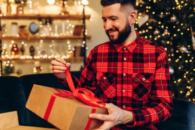 Happy young man opening Christmas gifts at home against the background of a decorated Christmas tree, Merry Christmas and New Year