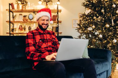 Happy young attractive man with a beard, wearing a Santa Claus hat, working with a laptop, sitting on a sofa in a Christmas house, against the background of a New Year tree
