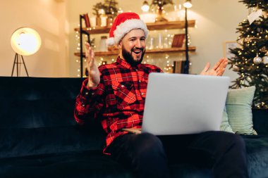 Excited young attractive man with a beard, wearing a Santa Claus hat, uses a laptop and rejoices, showing a victory gesture, sitting on the sofa at home against the background of a Christmas tree