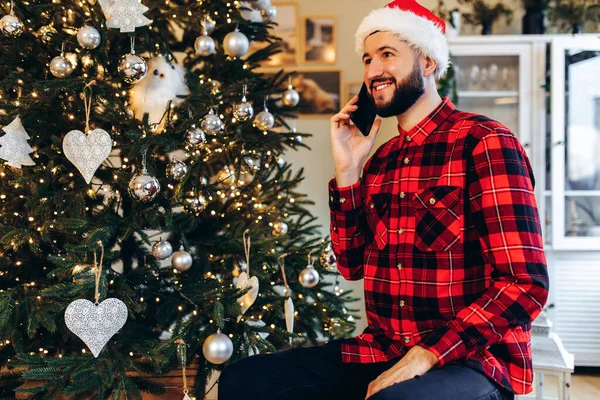 笑顔若いです男でサンタクロースの帽子話しています携帯電話にホームでクリスマスツリーの背景 — ストック写真