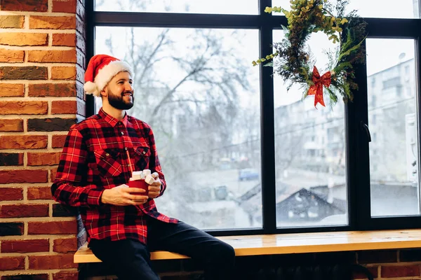 Attractive Christmas Man Wearing Santa Claus Hat Sits Windowsill Looks — Fotografia de Stock