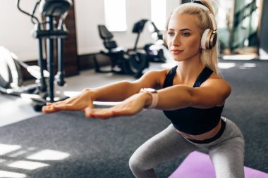 Fitness sporcusu genç bir kadın kulaklıkla müzik dinliyor ve spor salonunda, spor salonunda, sağlıklı bir yaşam tarzında çömelme yapıyor.