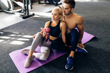 Spor kıyafetleri içinde yoga minderlerinin üzerinde oturan ve spor salonundaki antrenmandan sonra su şişelerini tutan genç bir çift.