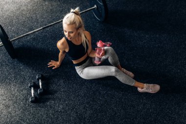 Spor giyim sektöründe genç bir kadın spor salonunda başarılı bir idmandan sonra elinde bir şişe suyla yerde oturuyor.