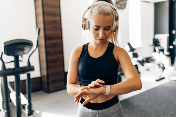 Çekici genç sporcu kadın spor salonuna gider, kulaklıkla müzik dinler ve elindeki spor saatine, sağlıklı yaşam tarzına bakar.