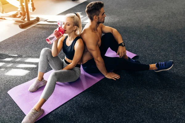 Spor kıyafetleri içinde yoga minderlerinin üzerinde oturan ve spor salonundaki antrenmandan sonra su şişelerini tutan genç bir çift.