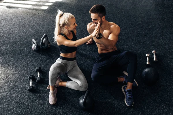 Sportif genç bir adam ve bir kadın spor salonunda yerde oturup birbirlerine beşlik çakıyorlar spor salonunda spor yaptıktan sonra spor salonundaki insanlara spor yapıyorlar.