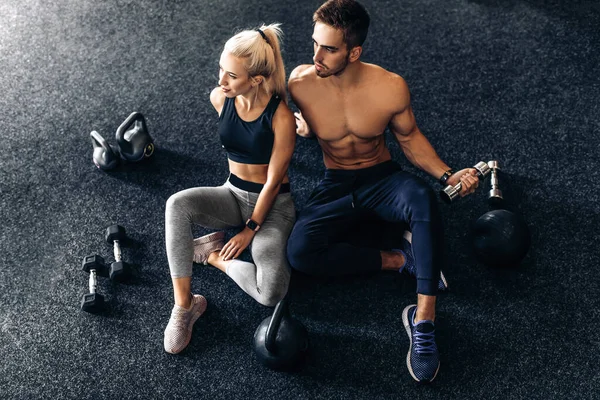 Young couple of athletic people, in sportswear, sitting together on the floor of the gym, with dumbbells, together after training in the gym