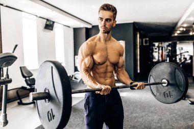 Genç kaslı bir adam spor salonunda halterle çalışıyor. Tişörtsüz atletik bir adam kol kaslarını çalıştırır ve halter egzersizi yapar.
