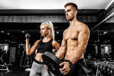 Genç, güzel, sportif bir kadın ve spor kıyafetleriyle spor salonunda halterleri kaldıran kaslı bir adam.