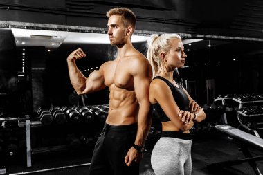 Güzel, sportif bir çift, siyah bir arka planda spor salonunda dururken kaslarını gösteriyor.