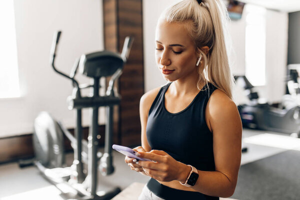 Красивая молодая фитнес-женщина, в спортивной одежде, расслабляется после тренировки использует мобильный телефон, в тренажерном зале