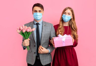 Sevgililer Günü kutlaması, birbirine aşık mutlu çift, yüzü tıbbi maskeli kadın ve erkek, virüsten korunmak için, izole edilmiş pembe arka planda bir buket çiçek ve hediyeyle