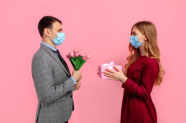Yakışıklı, şık bir adam yüzüne takım elbise ve tıbbi maske takıp güzel kız arkadaşına pembe arka planda çiçek ve hediye kutusu veriyor. Sevgililer günü konsepti, koronavirüs, karantina...