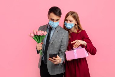 Tıbbi maskeli mutlu bir çift, kırmızı elbiseli bir kadın ve şık takım elbiseli bir adam, Sevgililer Günü 'nü kutluyorlar, kadınlar günü, cep telefonu kullanıyorlar ve pembe arka planda hediye kutusu tutuyorlar.