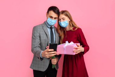 Tıbbi maskeli mutlu bir çift, kırmızı elbiseli bir kadın ve şık takım elbiseli bir adam, Sevgililer Günü 'nü kutluyorlar, kadınlar günü, cep telefonu kullanıyorlar ve pembe arka planda hediye kutusu tutuyorlar.