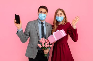 Sevgililer Günü 'nü hediye kutlayan tıbbi maskeler takan heyecanlı genç bir çift pembe arka planda cep telefonu kullanıyor.