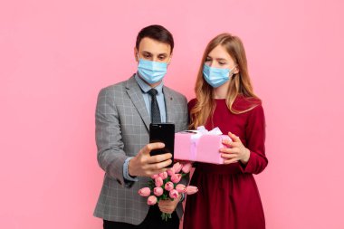 Tıbbi maskeli mutlu bir çift, kırmızı elbiseli bir kadın ve şık takım elbiseli bir adam, Sevgililer Günü 'nü kutluyorlar, kadınlar günü, cep telefonu kullanıyorlar ve pembe arka planda hediye kutusu tutuyorlar.