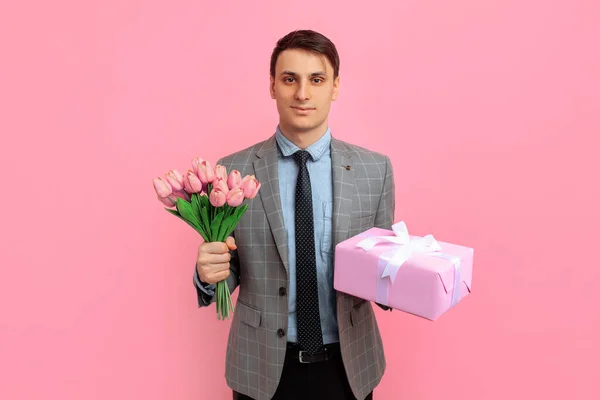 Elegant smiling man with gift box and bouquet of flowers, over isolated pink background, Concept for Valentine's Day, March 8