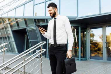İş adamı cep telefonu kullanıyor, ofisin dışındaki ofis binasının arka planında mesaj atıyor iş için elinde akıllı telefon tutan genç bir adam.